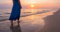 Woman in flowing blue dress walking barefoot on sandy beach at golden sunset with ocean waves and warm sky colors creating peaceful seaside moment.