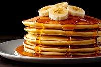 Stack of fluffy golden pancakes topped with fresh banana slices and dripping maple syrup on white plate against dark background, close up food photography.