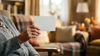 Senior woman holding blank white card in cozy living room with warm lighting, ideal for greeting cards, invitations, and promotional messaging.