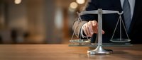 Businessman in suit touching scales of justice on wooden desk with blurred office background, representing law, legal services, and professional counsel.