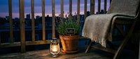 Cozy balcony at dusk with glowing lantern, lavender pot, and draped chair against a purple twilight city skyline backdrop.