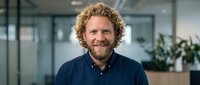 Smiling middle-aged Caucasian businessman with curly hair and beard in navy shirt standing in modern office workspace for corporate headshot photography.