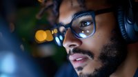 Young man with dark skin tone and beard wearing glasses and headphones, focused on screen work or gaming in low light environment with bokeh background.
