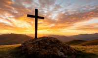 Christian cross silhouette on rocky hill against dramatic orange sunset sky and mountain landscape, spiritual symbol overlooking peaceful valley at dusk.