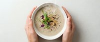 Female hands holding a creamy mushroom soup bowl garnished with fresh herbs and croutons, overhead view on white background for food photography and culinary content.