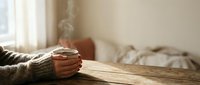 Cozy morning scene with hands holding warm coffee mug on wooden table in sunlit bedroom. Soft natural lighting creates peaceful atmosphere for relaxation.