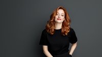Young woman with red wavy hair wearing black t-shirt smiling confidently against dark gray background, studio portrait in minimalist style for business and lifestyle content.