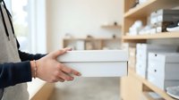 Person holding white box in modern kitchen with wooden shelves and storage containers, suitable for product packaging, delivery service, and home organization concepts.