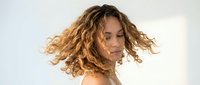Young mixed-race woman with curly blonde hair in motion against white background, expressing freedom and joy in natural light studio portrait.