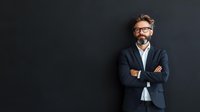 Confident mature Caucasian businessman with beard and glasses wearing navy blazer standing with arms crossed against dark wall, professional portrait.
