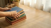 Woman's hands touching stack of colorful books on wooden table in bright living room with coffee cup and plants creating cozy reading atmosphere.