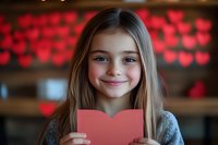 Young girl holding red heart card with blurred heart bokeh background, smiling warmly in gray sweater. Romantic holiday concept for Valentine's Day or Mother's Day.