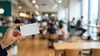 Woman holding blank white business card in modern office lobby with blurred colleagues and wooden interior, suitable for corporate branding and networking concepts.