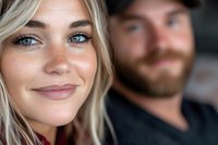 Close up portrait of young blonde woman with freckles and blue eyes smiling naturally, with bearded man in background. Soft focus romantic lifestyle photo.