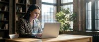 Professional Asian businesswoman working on laptop in modern office space with natural lighting and bookshelves for corporate presentations.