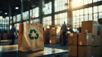 Paper shopping bag with green recycling symbol in warehouse or distribution center at sunset, cardboard boxes and workers in background, eco-friendly retail concept.
