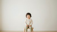 Young child with dark curly hair and light skin tone sitting on wooden stool against neutral wall background. Wearing cream-colored long-sleeve shirt. Suitable for parenting, childcare, education, and family-related design projects.