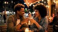 Young African American couple enjoying romantic dinner date at outdoor restaurant patio with string lights, toasting with wine glasses in evening atmosphere.