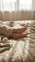 Bare feet resting on soft linen bedding in warm morning light, with a mug nearby, evoking comfort, relaxation, and a cozy lazy weekend.