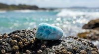 Larimar gemstone on volcanic rock by turquoise ocean waves. Natural healing crystal mineral specimen against tropical seascape background for wellness and spirituality.