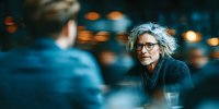 Senior woman with gray hair and glasses having conversation in modern cafe with warm bokeh lighting background for lifestyle and communication concepts.