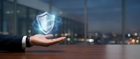 Businessman hand holding glowing digital shield icon representing cybersecurity, data protection and business security solutions in modern office.