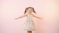 Happy young girl with pigtails dancing in floral dress against pink gradient background. Joyful child with arms outstretched expressing freedom and happiness.