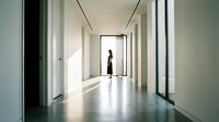 Woman in dark clothing standing in modern minimalist hallway with polished concrete floor, white walls, and natural light streaming through large windows, suitable for corporate, architectural, and lifestyle design projects.