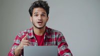 Young Hispanic man in plaid shirt expressing surprise while working on laptop, gesturing with raised finger against gray background. Professional workplace portrait.
