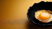 Fresh sunny side up egg with crispy golden edges served in rustic black ceramic bowl against warm brown background, minimalist food composition.
