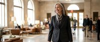 Professional middle-aged Caucasian businesswoman in dark suit walking confidently through modern office lobby with arched windows and contemporary furniture.
