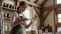Woman in apron sifting flour into bowl in rustic kitchen with wooden beams and shelves of jars, baking preparation.