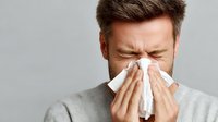 Caucasian man with closed eyes blowing nose into tissue against gray background, showing symptoms of cold, flu, or allergies.