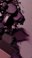 Crushed purple eyeshadow palette with scattered powder texture and metallic shimmer creating abstract patterns on pink background. Macro cosmetic photography.