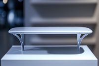 Minimalist curved shelf in light gray with elegant support brackets mounted on white surface against blurred background, modern interior design element.