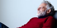 Senior Caucasian man with gray beard wearing red sweater laughing heartily while relaxing in chair, expressing genuine joy and happiness against light background.