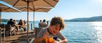 Young man enjoying refreshing drink at lakeside restaurant terrace during summer vacation with mountain views and sparkling water reflections.