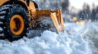 Snow removal equipment clearing winter road during snowfall with bokeh lights in background, close up view of industrial wheel loader bucket.