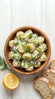 Potato salad with creamy dressing, fresh dill, red onion, and herbs served in wooden bowl with lemon and bread on white wooden surface.