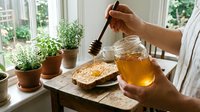 Person drizzling honey onto toast at bright windowsill with potted herbs and glass jar, ideal for food and lifestyle content.