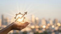 Businessperson holding glowing gear and upward arrow icon against blurred cityscape at sunset, representing business growth, productivity improvement, and success strategy.