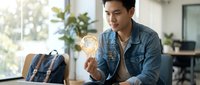 Young Asian man in denim jacket holding glowing light bulb representing creative ideas and innovation in modern office workspace for business concepts.
