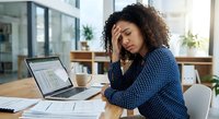 Stressed African American businesswoman with curly hair experiencing headache while working on laptop with spreadsheets in modern office workspace.