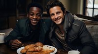 Two diverse friends enjoying dinner together at home, sharing bread and conversation in warm evening lighting, creating authentic social connection moment.