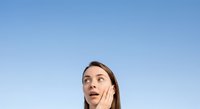 Young woman with brown hair and fair complexion, appearing surprised or amazed, hand touching face against clear blue sky background for advertising and marketing campaigns.