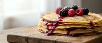 Stack of golden pancakes topped with fresh raspberries and blackberries, berry syrup dripping down sides on rustic wooden cutting board in bright kitchen.