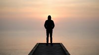 Silhouetted male figure standing on wooden pier at sunrise or sunset, gazing toward calm water horizon. Contemplative scene suitable for motivational, inspirational, and lifestyle content.