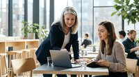 Two professional businesswomen collaborating on laptop in modern office workspace. Female colleagues discussing project at wooden table with plants.
