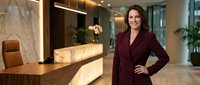 Professional businesswoman in burgundy suit standing confidently in modern corporate office lobby with marble reception desk and contemporary lighting.