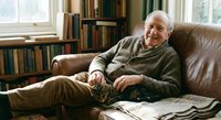 Elderly Caucasian man relaxing in leather armchair at home library, wearing green sweater, surrounded by books and newspapers in cozy reading room.